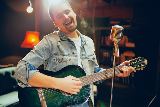 Talented bearded Caucasian musician playing acoustic guitar and singing at home studio.