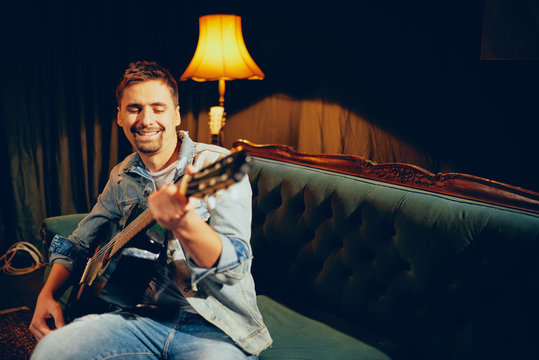 Caucasian talented bearded musician playing his acoustic guitar and sitting on sofa. Living room interior.