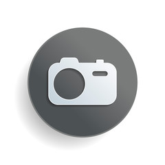 Simple photo camera. Technology icon. White paper symbol on gray round button or badge with shadow