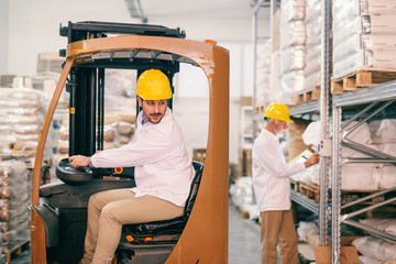Young warehouse worker driving forklift. In background senior worker with helmet on head checking...