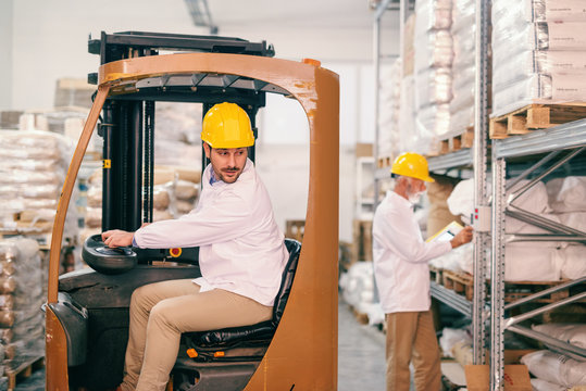 Young warehouse worker driving forklift. In background senior worker with helmet on head checking temperature in warehouse.