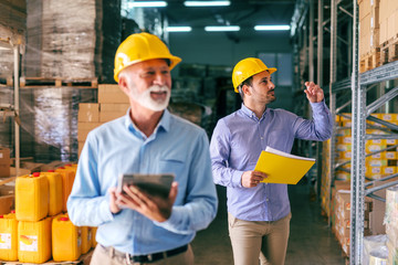 Two businessmen in warehouse. Selective focus on younger one with helmet on head and folder with documents in hand.