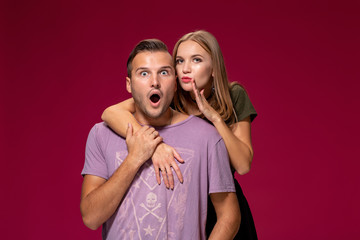 Studio shot of secret young woman and man keep index fingers on lips, stand next to each other, tell private information, isolated over burgundy background.
