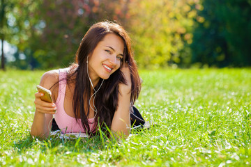 Naklejka na ściany i meble Young woman in dress lies on green grass and listens to music in headphones through smartphone.