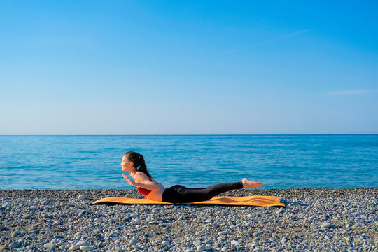 Young slim woman in tight sportswear doing locust pose on orange yoga mat outdoors at pebble beach by the sea. Yoga at nature concept
