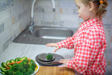 Cute baby girl in shirt is cutting with a knife vegetables cabbage on a wooden board next to...
