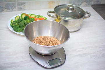 raw buckwheat and fresh vegetables on the kitchen table. Weighing cereals on kitchen scales. Healthy eating concept