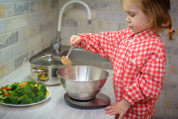 cute baby weighs buckwheat and vegetables on kitchen scales. A little girl is cooking vegetarian...