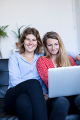 Image of two smiling young woman sitting on the sofa with laptop.