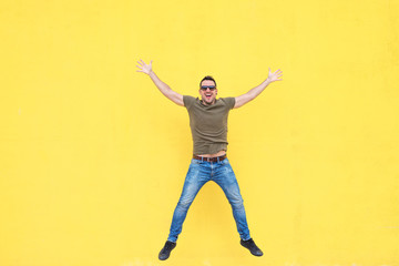Fototapeta na wymiar Young man with sunglasses jumping against a yellow wall.