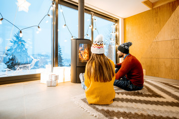 Young couple dressed in bright sweaters sitting near the fireplace in the modern house in the mountains durnig winter time - obrazy, fototapety, plakaty