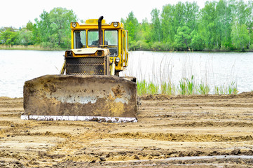 Yellow crawler tractor clears the construction site for recreation on the lake. - obrazy, fototapety, plakaty