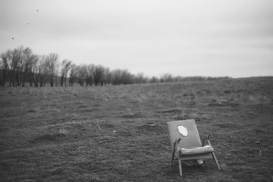 A hill with trees in black and white. Vintage armchair in the middle of the field. Gray landscape of nature and old furniture. Armchair with wooden handles. Autumn bare trees. Dry grass on the field.