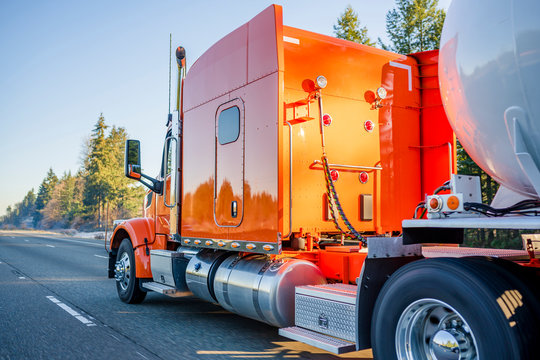 Bright orange big rig semi truck transporting tank semi trailer for transportation of liquid and liquefied chemical cargo running on the road