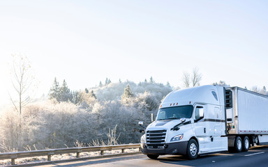 Bright white comfort ergonomic big rig semi truck with refregerated semi trailer running on winter highway wit frost hill