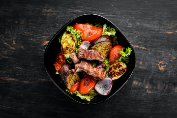 Baked beef and grilled vegetables in a black plate. On a wooden background. Top view. Free space for your text.