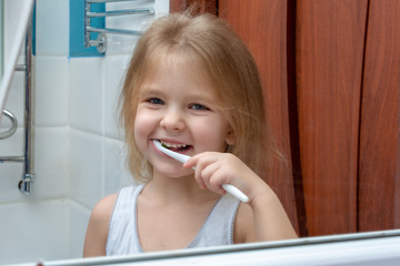 A little girl with blond hair brushing her teeth. The child is smiling at the reflection in the mirror.