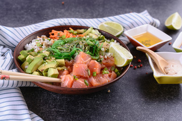 Homemade healthy Poké bowl with rice, salmon, avocado, white onion, scallion greens, sesame oil, soy and sesame sauce, seaweed (wakame) and other ingredients on a dark backgound are ready to eat