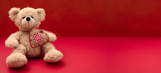 Valentines day. Teddy bear with a fabric heart against red background, banner, copy space