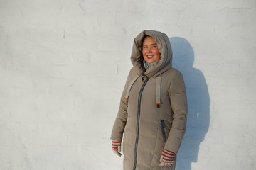 Portrait of pretty cheerful rosy girl clothed in winter jacket with hood at white stone wall background in sunny winter day.