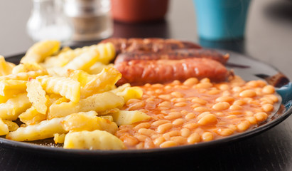 A close-up image of sausage, chips and baked beans on a black plate