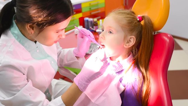 children's dentist examines the teeth and mouth of a cute little red-haired girl in a yellow-red dental chair. Pediatric dentistry