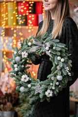 Christmas shopping. Green Christmas wreath in female hands. Decorated with gray elements, cotton and silver ball . Garland bokeh on background.