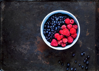 Raspberries and bilberry in  blue bowl  on vintage rusty metal background. Berry picking, organic food concept. 