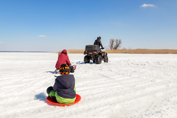 Man on ATV quadbike riding sledges with kids in tow on frozen lake surface at winter. Winter extreme sports and recreation. Children outdoor fun and activities