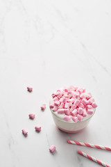 Hot chocolate or cocoa with whipped cream and pink marshmallow candy in pink and white mug with pink striped straws on white marble table. Copy space for text.