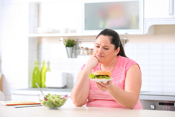 Sad overweight woman choosing between salad and burger in kitchen. Healthy diet