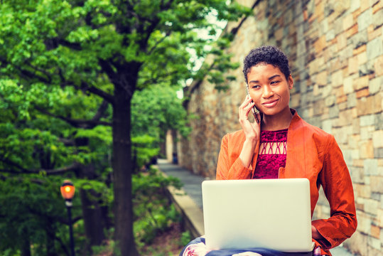 Young African American Woman studying, working on laptop computer, talking on cell phone outside in New York City, wearing orange red jacket, flower patterned skit, sitting by trees, walls on campus..