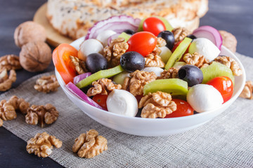 Salad with cherry tomatoes, mozzarella cheese, black olives, kiwi, and walnuts on black wooden background.