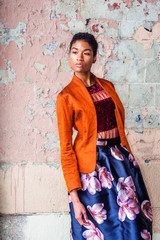 Portrait of Young African American Woman in New York City, with short afro hair, wearing fashionable orange red jacket, dark red under top, dark blue flower patterned skirt, standing by painted wall..