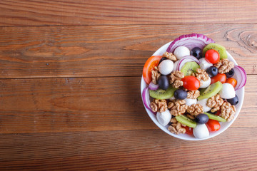 Salad with cherry tomatoes, mozzarella cheese, olives, kiwi, and walnuts on brown wooden background.