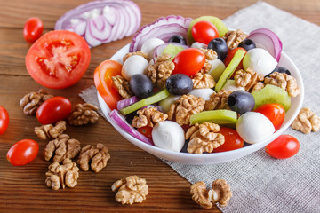 Salad with cherry tomatoes, mozzarella cheese, olives, kiwi, and walnuts on brown wooden background.