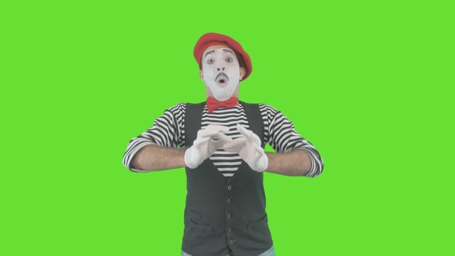 Pantomime man with facial paint posing for camera blowing up balloon, isolated on green background