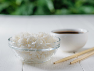Glass bowl with white long grain rice, bowl with soy sauce and wooden sticks on white table on green tree background.