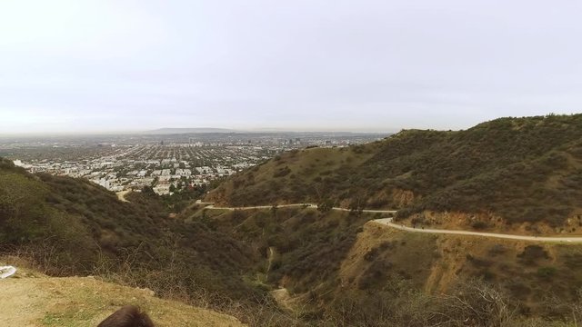 A dolly up shot behind a sitting hiker revealing Runyon Canyon on an overcast day. Hikers on the trail in the distance.  	