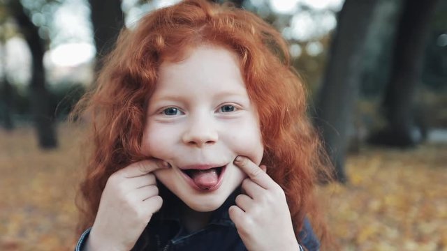 close up portrait of cute little redhead girl standing in autumn park. Funny baby girl showing grimaces and laughing