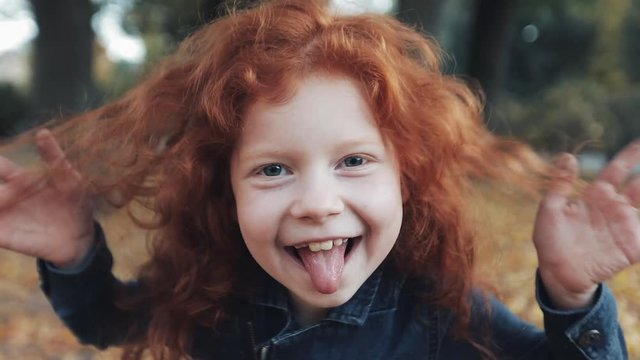 close up portrait of cute little redhead girl standing in autumn park. Funny baby girl showing grimaces and laughing