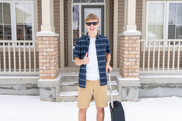 Excited young traveler with his suitcase leaving his home for a winter vacation. He is wearing shorts and a t shirt while standing in the snow.