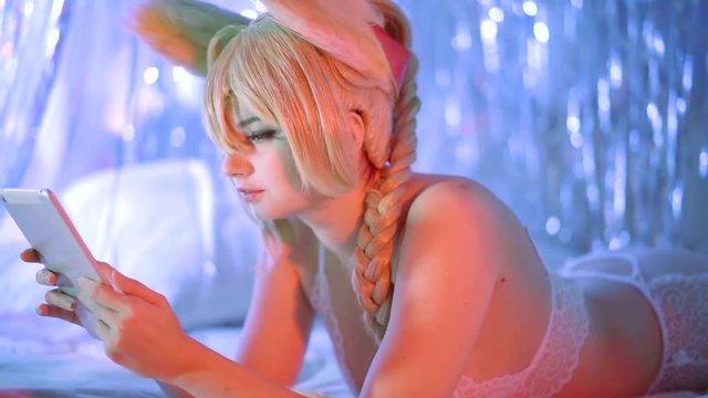 Side view shot of a young charming girl sitting online on a tablet and swinging her legs lying in bed in sexy white lingerie.
