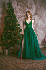 Obraz na płótnie Canvas Winter holidays, celebration and people concept - young sexy woman in elegant green evening dress over christmas interior background