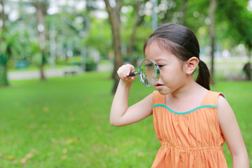 Cute little Asian child girl looking through magnifiying glass on at grass outdoors.