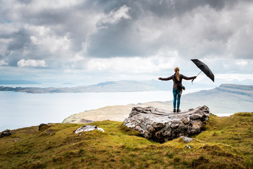 Woman stands on big rock with arms wide open and an umbrella in her hand, rainy weather in the scottish highlands, light and hope, after the rain