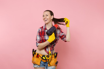 Young beautiful fun handyman woman in plaid shirt, denim shorts, kit tools belt full of variety instruments holding power heat gun isolated on pink background. Female in male work. Renovation concept