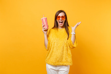 Happy young woman in heart orange glasses spreading hands, screaming, holding plastic cup with cola or soda isolated on yellow background. People sincere emotions, lifestyle concept. Advertising area.