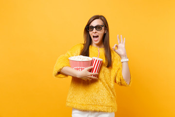 Amazed young woman in 3d imax glasses watching movie film, holding bucket of popcorn, cup of cola or soda, showing OK sign isolated on yellow background. People sincere emotions in cinema, lifestyle.