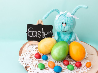 Easter celebration. Cute handmade knitted hare - a symbol of the holiday. Near her are colored eggs and Easter cake. Free space for text. Greeting card.